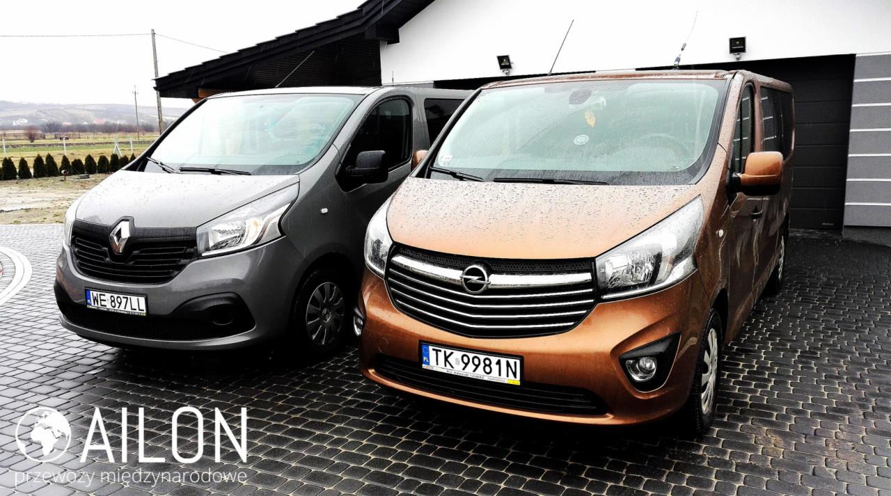 Dwa busy: szary Renault Trafic i pomarańczowy Opel Vivaro, zaparkowane na brukowanej nawierzchni przed budynkiem w deszczowy dzień.