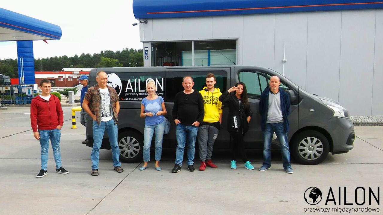 Grupa osób stoi przed szarą furgonetką z logo firmy przewozowej na tle stacji benzynowej.