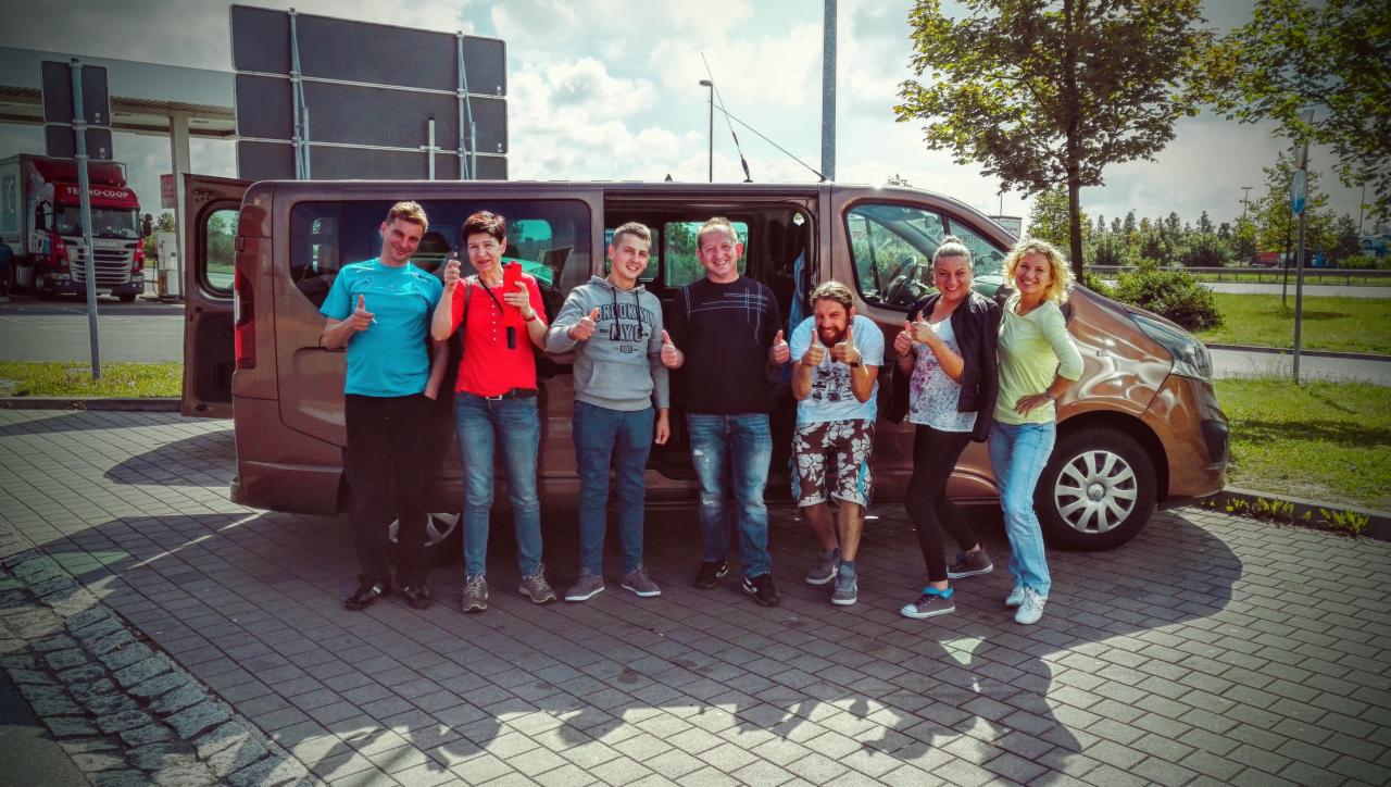 Grupa siedmiu osób, w tym jedna z brodą, pozuje z podniesionymi kciukami przed brązowym vanem z otwartymi drzwiami na parkingu z kostki brukowej, w tle stacja benzynowa i ciężarówka.