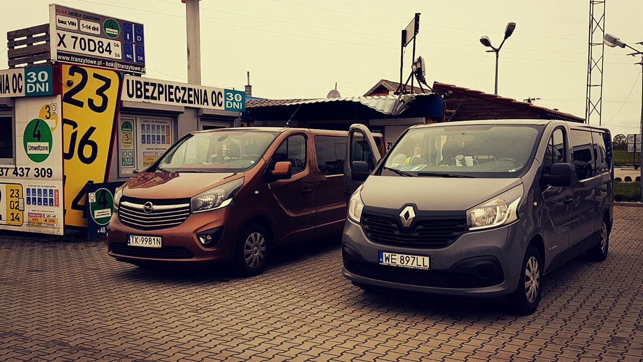 Dwa busy: brązowy Opel Vivaro i szary Renault Trafic zaparkowane na kostce brukowej przed budką z ubezpieczeniami OC i tablicami tranzytowymi, widoczne numery rejestracyjne.