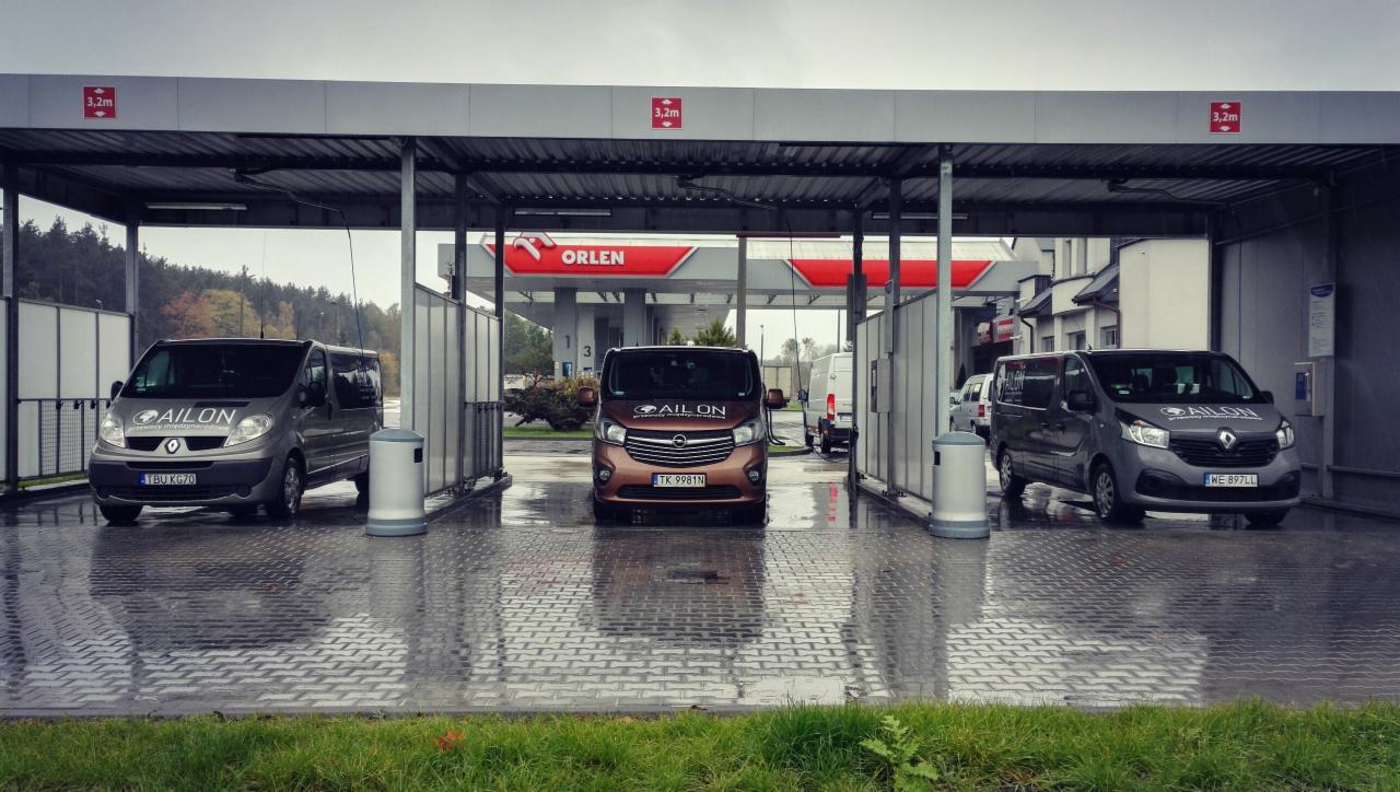 Trzy busy firmy Ailon zaparkowane pod zadaszeniem myjni samochodowej, w tle stacja Orlen.