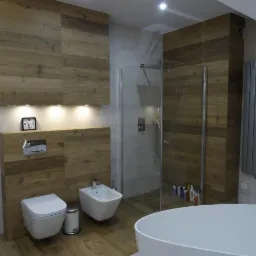 Nowoczesna łazienka z drewnianymi panelami ściennymi, podświetlanymi półkami, wiszącą toaletą i bidetem, kabiną prysznicową typu walk-in oraz wolnostojącą wanną z zaokrąglonymi krawędziami.