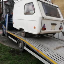 DroCar - Laweta przewozi białą, nieco zabrudzoną przyczepę kempingową na tle miejskiej zabudowy w pochmurny dzień.