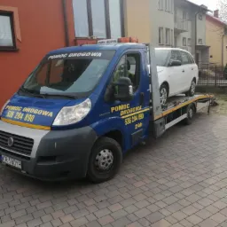 Niebieska laweta z białym samochodem osobowym na platformie, zaparkowana na brukowanej posesji przed budynkami mieszkalnymi w dzień.