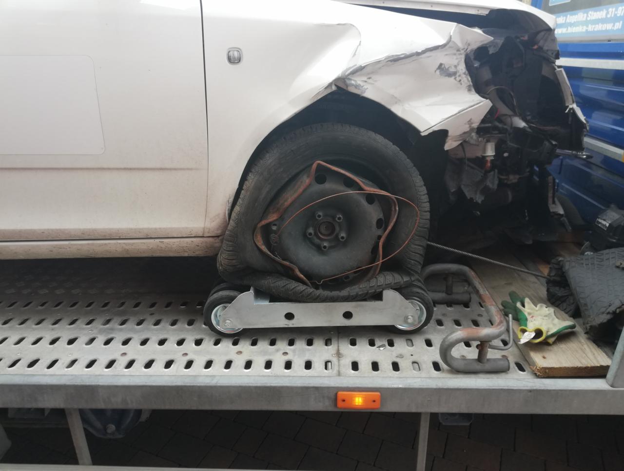 Uszkodzony biały samochód z przebitą oponą, umieszczony na platformie z kółkami na lawecie, widoczne uszkodzenia karoserii.