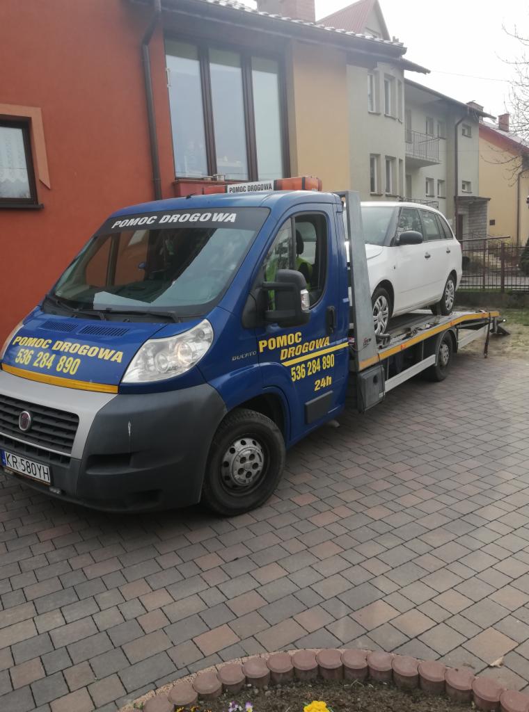 Niebieska laweta z białym samochodem osobowym na platformie, zaparkowana na brukowanej posesji przed budynkami mieszkalnymi w dzień.