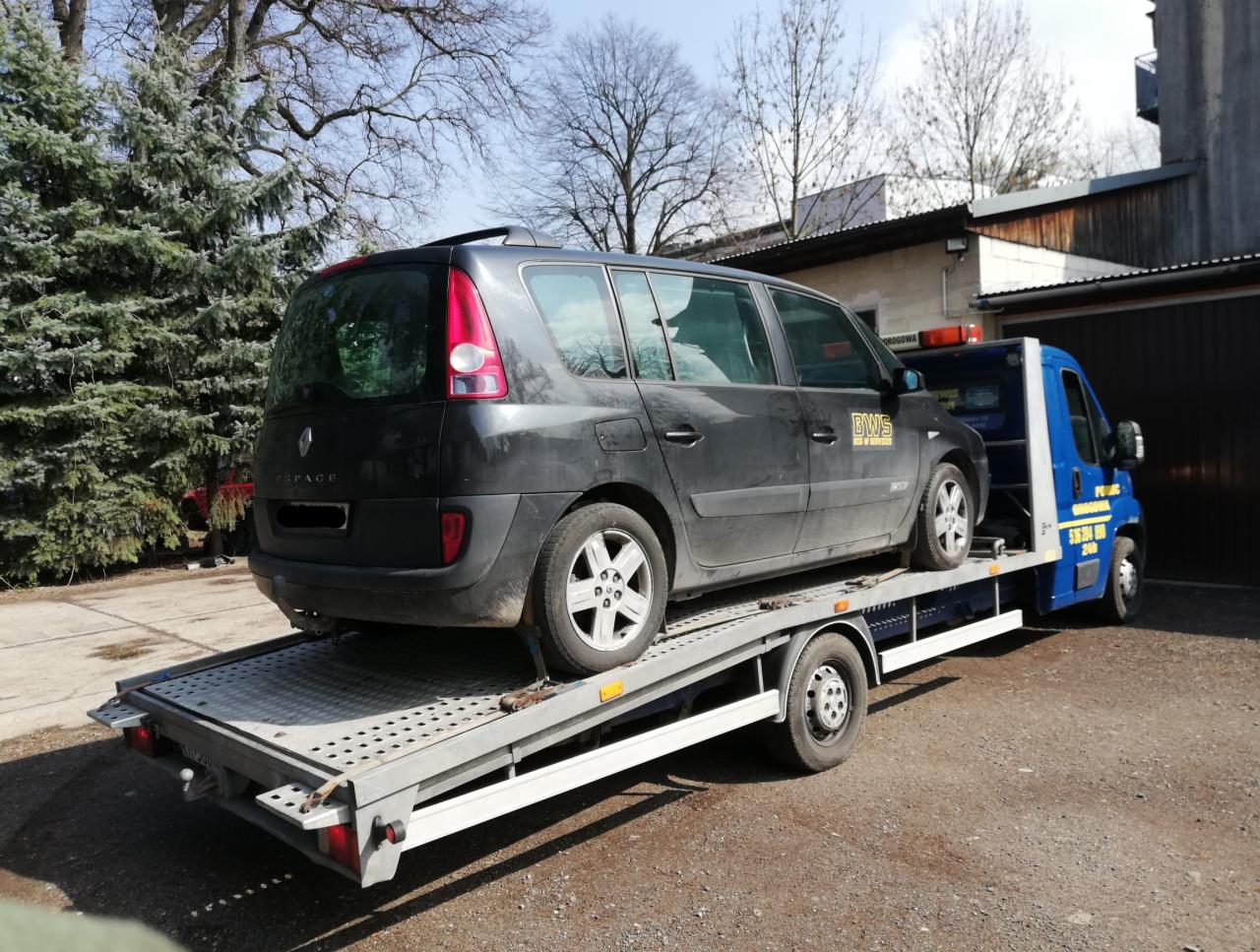 Czarny samochód Renault Espace na platformie lawety w trakcie załadunku lub transportu, widoczny fragment niebieskiej kabiny lawety z napisem reklamowym.