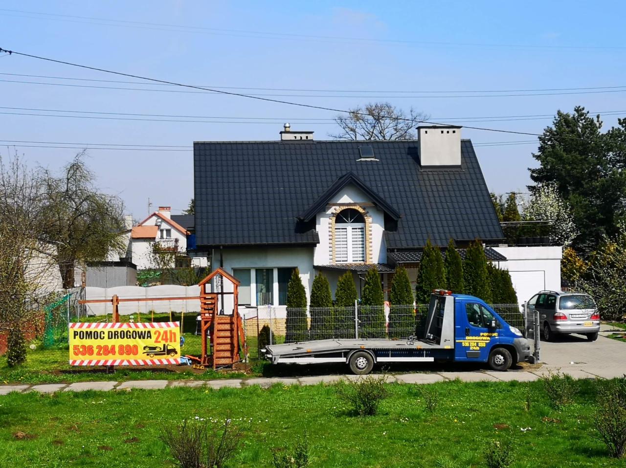 Niebieska laweta z zaparkowanym za nią srebrnym samochodem osobowym na tle domu jednorodzinnego w Krakowie; na pierwszym planie żółty baner z napisem 'Pomoc Drogowa 24h'.