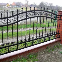 Eleganckie ogrodzenie kute z ceglanymi słupkami i ozdobnymi detalami, na tle zielonego trawnika i zabudowań mieszkalnych. Czarne elementy metalowe z subtelnymi zdobieniami.