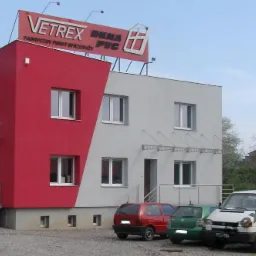 Dwukondygnacyjny budynek z czerwono-szarą elewacją, z oknami PCV, z szyldem reklamowym na dachu, oraz zaparkowanymi samochodami osobowymi i busem z logo firmy.