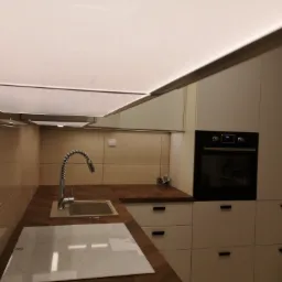 Nowoczesna kuchnia IKEA z drewnianym blatem, białymi szafkami i płytą indukcyjną, oświetleniem sufitowym LED i chromowaną baterią kuchenną.