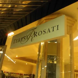 Podświetlany szyld sklepowy z nazwą 'Teresa Rosati' w eleganckim stylu, widoczny z dołu, z odblaskami na suficie i częściowo widocznym szyldem 'Liberty' obok.