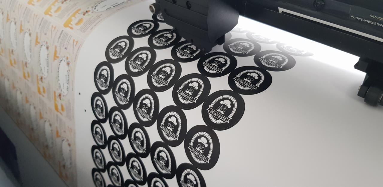 Proces produkcji okrągłych, czarno-białych naklejek z logo 'Urban Bikes' na arkuszu w drukarce wielkoformatowej.