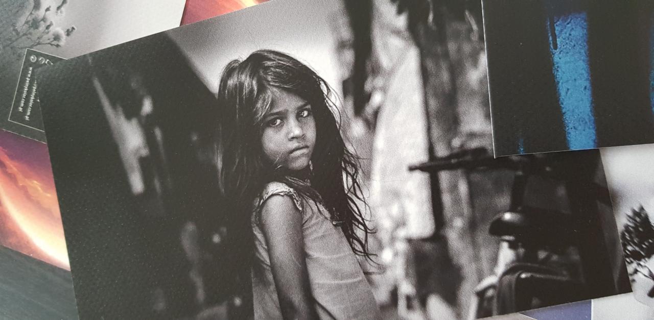 Stos wydrukowanych fotografii, w tym czarno-białe portretowe zdjęcie dziewczynki na pierwszym planie, z widocznymi innymi kartkami z różnymi grafikami w tle.