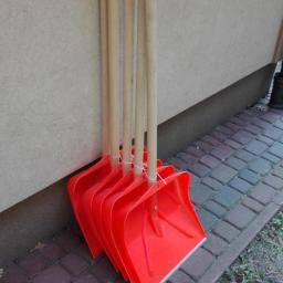 SKOL-SERVICES - Stojące przy ścianie cztery nowe, czerwone łopaty do śniegu z drewnianymi trzonkami, związane sznurkiem.