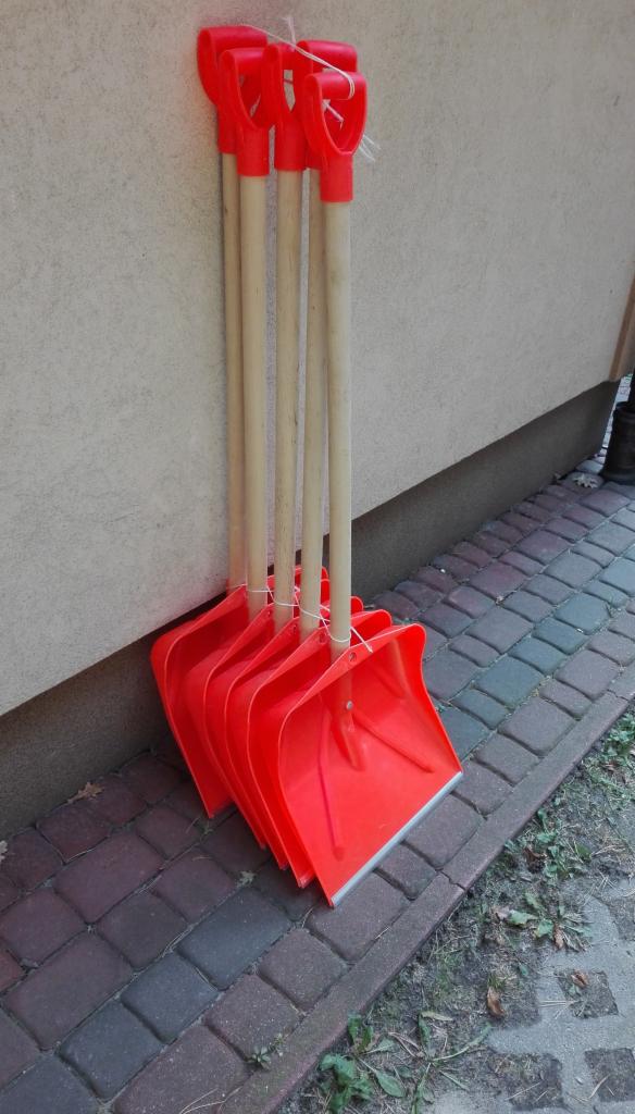 Stojące przy ścianie cztery nowe, czerwone łopaty do śniegu z drewnianymi trzonkami, związane sznurkiem.