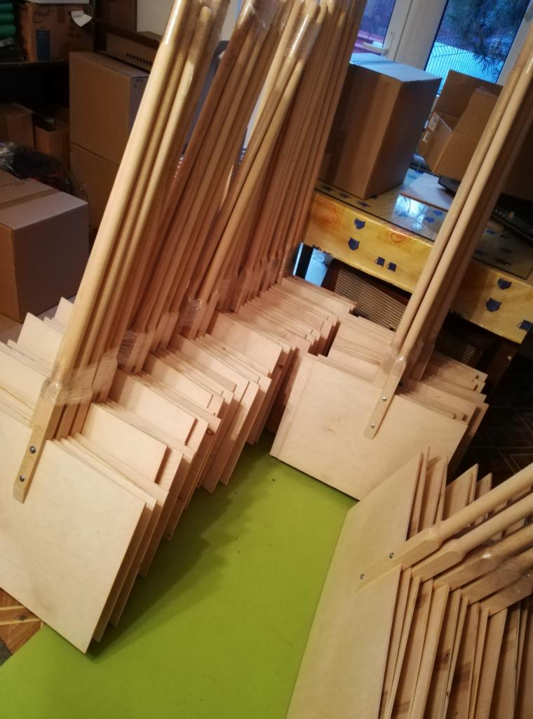 Stos drewnianych łopat do śniegu z długimi trzonkami, ułożonych rzędami na zielonej powierzchni, w tle kartony i stół.