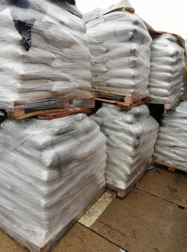 Stosy worków z jasną zawartością, owiniętych folią i ułożonych na drewnianych paletach, składowane na zewnątrz.