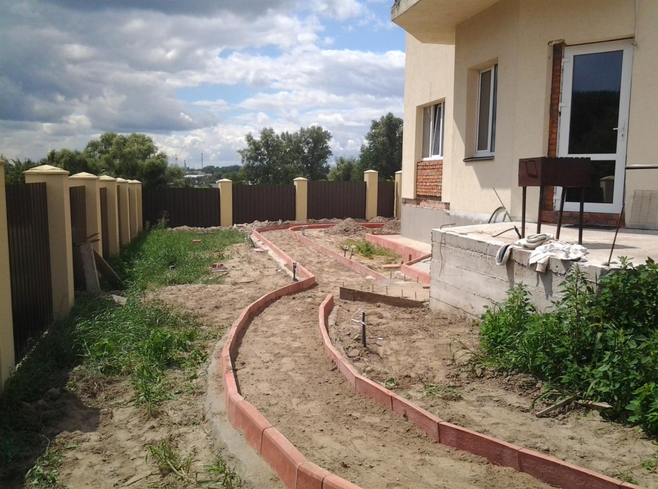 Kształtowanie ścieżki z obrzeży betonowych w ogrodzie, widoczne wystające rurki instalacji nawadniającej i fragment budynku w tle.