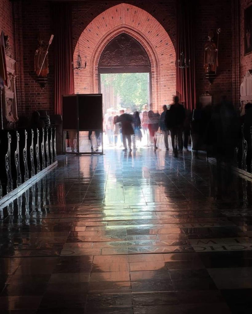 Wnętrze kościoła z ceglanymi ścianami i wysokim łukowym wejściem, przez które widać rozmyte sylwetki ludzi w ruchu, odbijające się w błyszczącej posadzce.