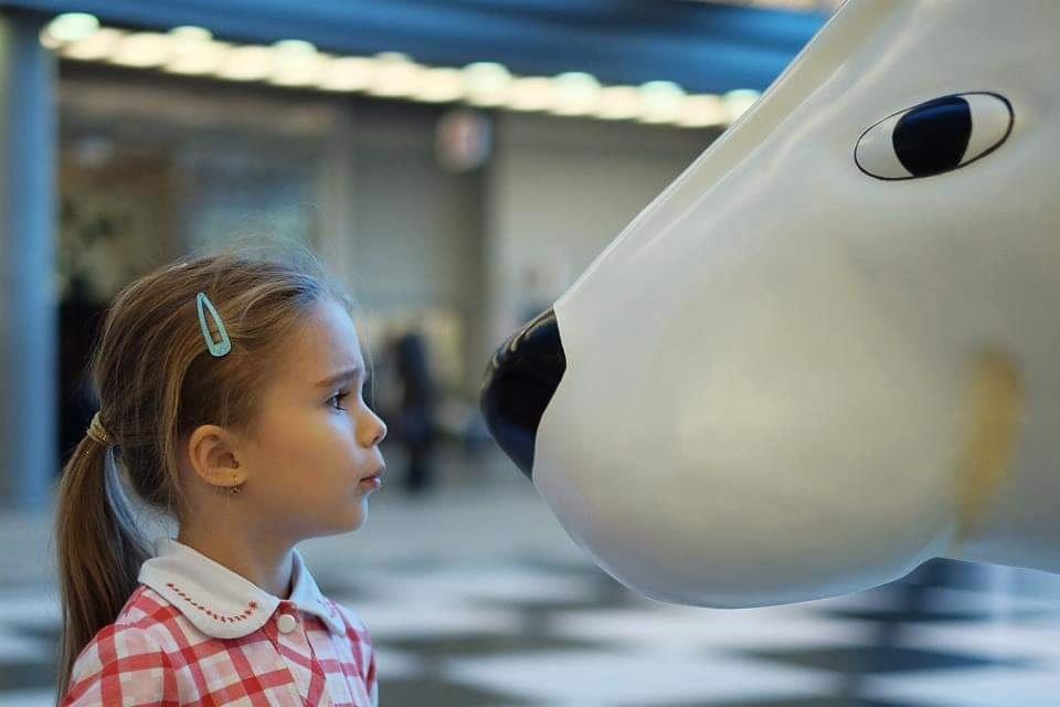 Dziewczynka z blond włosami i niebieską spinką, w czerwono-białej koszuli w kratę, patrzy z zaciekawieniem na ogromny, biały nos rzeźby niedźwiedzia polarnego.