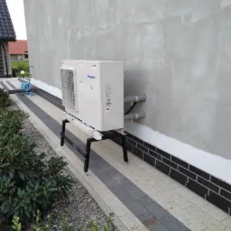 Zewnętrzna jednostka pompy ciepła Panasonic zamontowana na czarnym stelażu przy szarej ścianie domu z widocznymi rurami instalacyjnymi i kostką brukową.