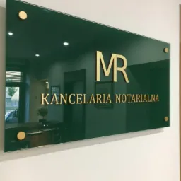 Szklana tablica informacyjna z napisem 'MR Kancelaria Notarialna' w złotym kolorze, zamocowana na ścianie za pomocą złotych śrub dystansowych, odbijająca wnętrze kancelarii z recepcją i oknem.