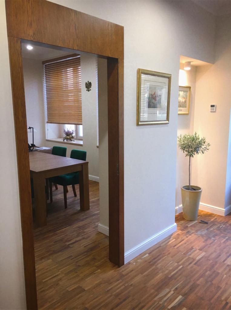 Wnętrze kancelarii z drewnianą podłogą parkietową, widok na stół i krzesła przez drewniane obramowanie drzwi, na ścianie obraz w złotej ramie i drzewko oliwne w doniczce.