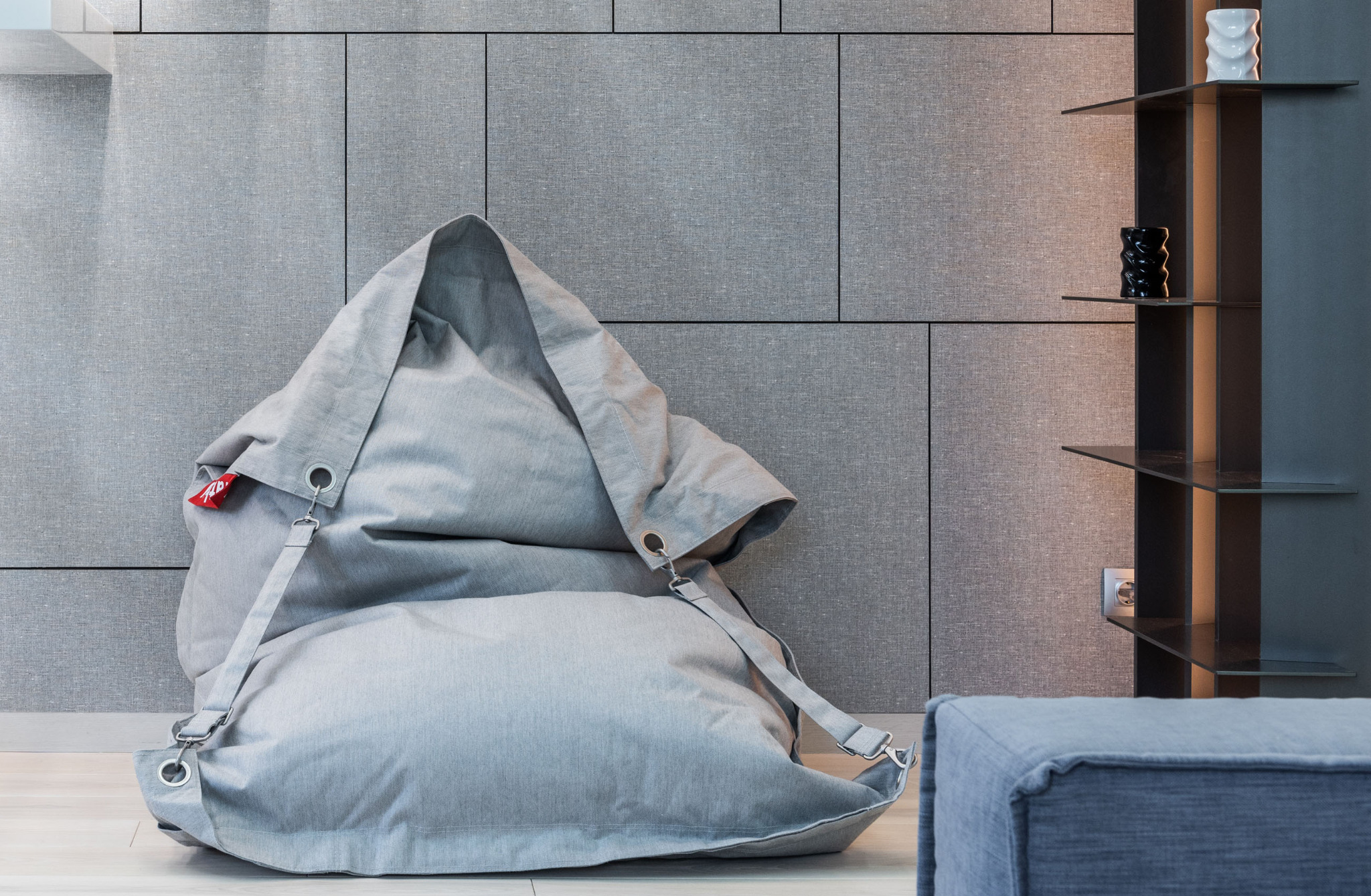 Szary puf w kształcie worka z metalowymi elementami i czerwoną metką na tle ściany z paneli tekstylnych i regału z dekoracjami.