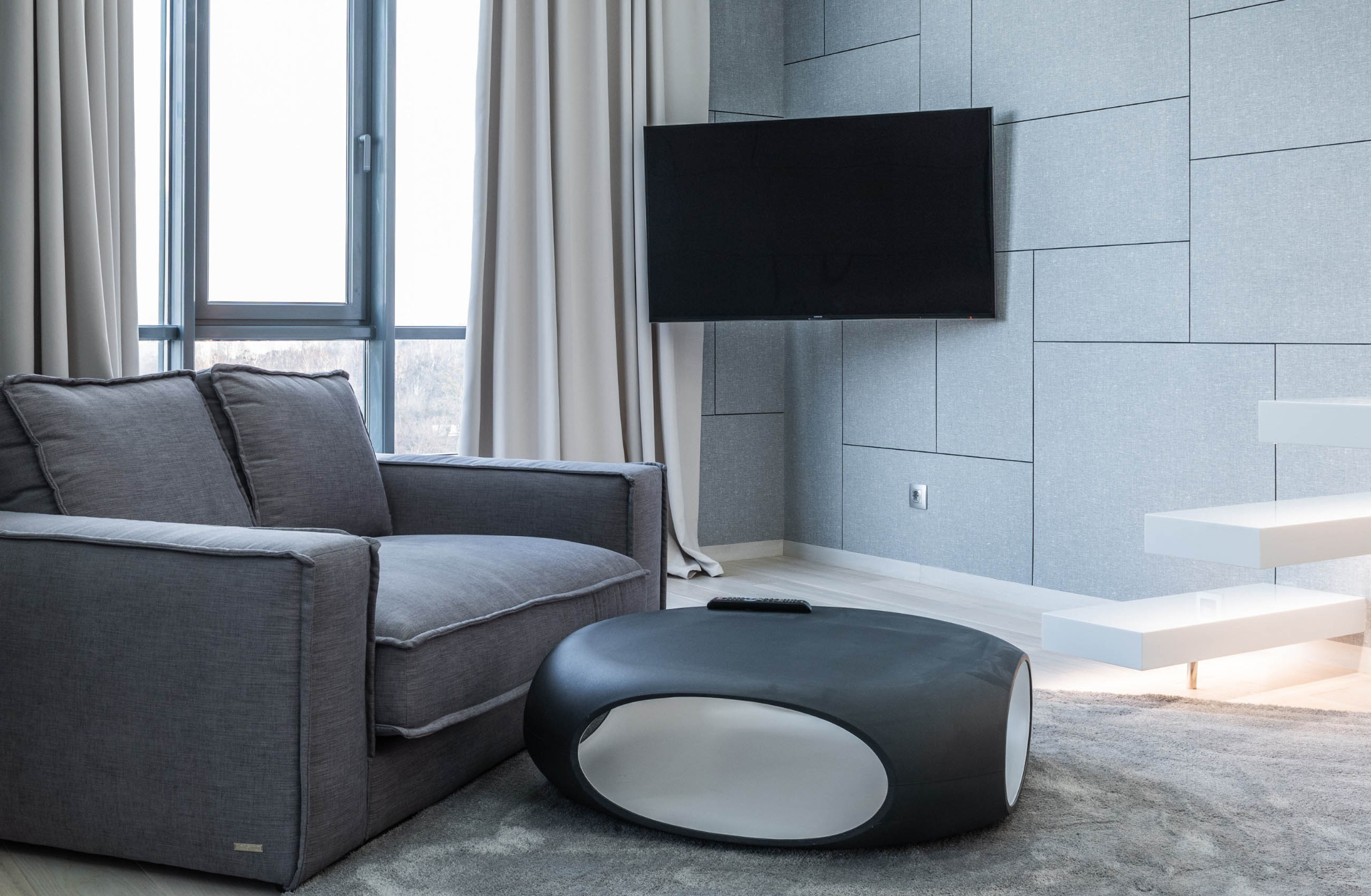 Szary narożnik z poduszkami w nowoczesnym salonie z telewizorem na ścianie z paneli, okrągły stolik kawowy z wycięciem i minimalistyczne białe schody.