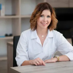 Uśmiechnięta kobieta w białym fartuchu siedzi za biurkiem w gabinecie, w tle regał z książkami i czarne krzesło biurowe.