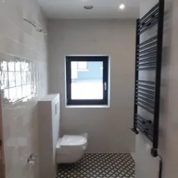 Wykończona łazienka z białymi, błyszczącymi płytkami ściennymi, wzorzystą podłogą, wiszącą toaletą z zabudowaną spłuczką i czarnym grzejnikiem drabinkowym, małe okno z czarną ramą.