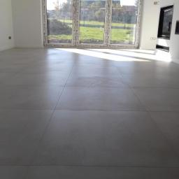 DARMI REMONTY I WYKOŃCZENIA WNĘTRZ - Duże, szare płytki podłogowe w jasnym pokoju z dużym oknem i widokiem na zielony trawnik, z fragmentem pomarańczowego ubrania po prawej stronie kadru.