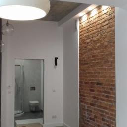 DARMI REMONTY I WYKOŃCZENIA WNĘTRZ - Wnętrze po remoncie: białe ściany, dekoracyjna ściana z czerwonej cegły, nowoczesne oświetlenie (lampa sufitowa i kinkiety), widoczny fragment łazienki z kabiną prysznicową, pracownik...