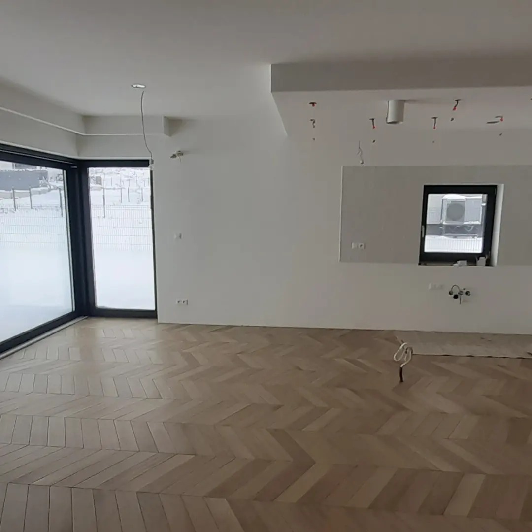 Jasne, przestronne wnętrze z parkietem ułożonym we wzór jodełki, duże okna z czarnymi ramami i przygotowane punkty oświetleniowe na suficie.