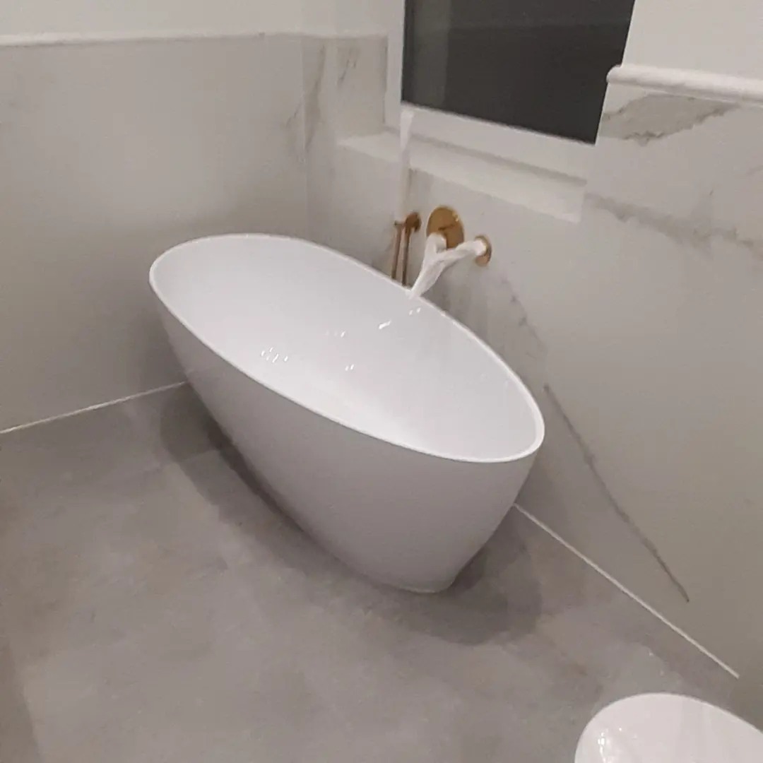 Wolnostojąca, owalna wanna w nowoczesnej łazience z szarymi płytkami podłogowymi i marmurowymi płytkami ściennymi, złota bateria ścienna.