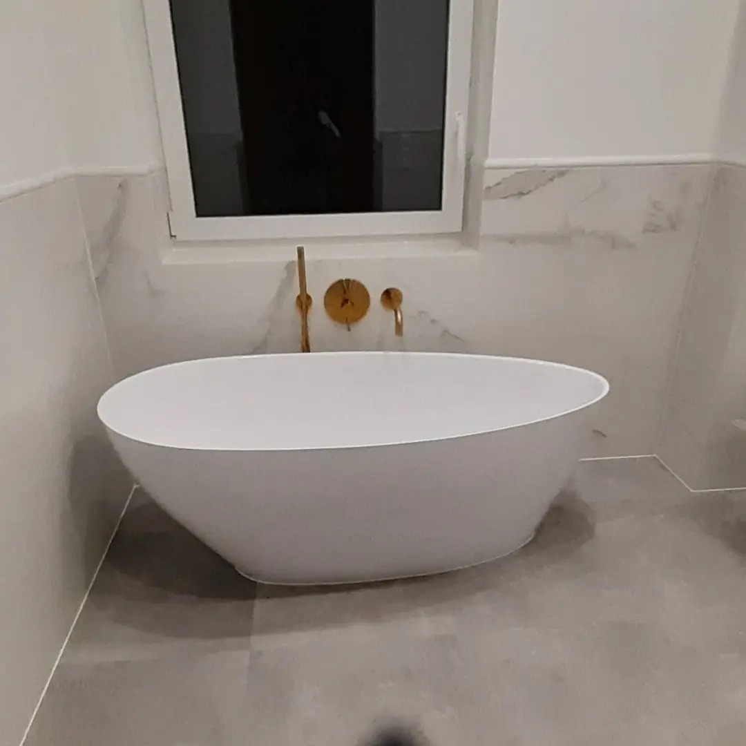 Wolnostojąca wanna owalna z baterią podtynkową w kolorze złotym na tle marmurowych płytek w łazience po remoncie.