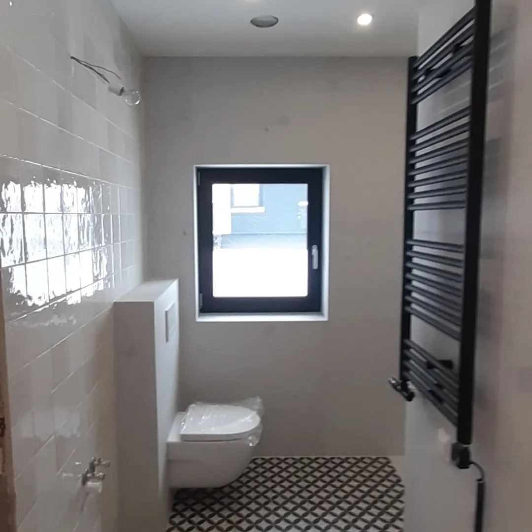 Wykończona łazienka z białymi, błyszczącymi płytkami ściennymi, wzorzystą podłogą, wiszącą toaletą z zabudowaną spłuczką i czarnym grzejnikiem drabinkowym, małe okno z czarną ramą.