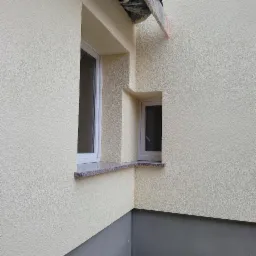 Fragment elewacji budynku po ociepleniu, z widocznymi dwoma oknami o różnej wielkości i granitowymi parapetami, dolna część ściany pomalowana na szaro.