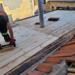 VERTEX EXPERT SPÓŁKA Z OGRANICZONĄ ODPOWIEDZIALNOŚCIĄ - Pracownik w czerwonych rękawicach montuje drewniane deski na dachu, widoczne narzędzia i fragment starej dachówki.