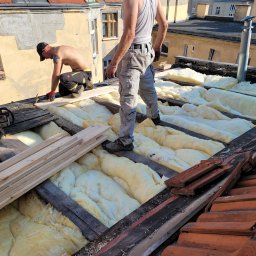 VERTEX EXPERT SPÓŁKA Z OGRANICZONĄ ODPOWIEDZIALNOŚCIĄ - Prace dekarskie: ocieplanie dachu wełną mineralną, widoczni dekarze w trakcie montażu, drewniane elementy konstrukcji dachowej, częściowo zdjęte dachówki.