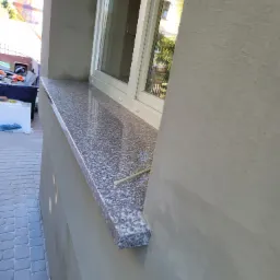 Zewnętrzny parapet z szarego granitu z widoczną fakturą, zamontowany pod oknem w budynku z nową elewacją, na pierwszym planie fragment chodnika z kostki brukowej.