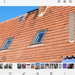 VERTEX EXPERT SPÓŁKA Z OGRANICZONĄ ODPOWIEDZIALNOŚCIĄ - Czerwona dachówka z trzema oknami dachowymi i ceglanym kominem na tle błękitnego nieba. Widoczne ślady użytkowania na dachówkach.