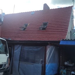 VERTEX EXPERT SPÓŁKA Z OGRANICZONĄ ODPOWIEDZIALNOŚCIĄ - Czerwony dach domu z oknami dachowymi, widziany z dołu. Na dachu pracownik w podnośniku koszowym. Częściowo widoczny biały samochód dostawczy.