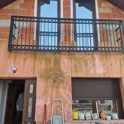 VERTEX EXPERT SPÓŁKA Z OGRANICZONĄ ODPOWIEDZIALNOŚCIĄ - Dom w trakcie ocieplania: widoczna warstwa izolacji z kołkami, balkon z czarną balustradą, okna z roletami i drabina oparta o ścianę. Częściowo widoczne wiadra z farbą.