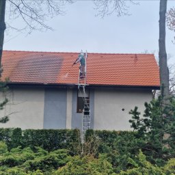 VERTEX EXPERT SPÓŁKA Z OGRANICZONĄ ODPOWIEDZIALNOŚCIĄ - Pracownik na drabinie czyści dach z czerwonej dachówki za pomocą myjki ciśnieniowej. Widoczny budynek z szarą elewacją i zielone krzewy w tle.