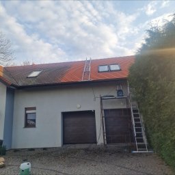 VERTEX EXPERT SPÓŁKA Z OGRANICZONĄ ODPOWIEDZIALNOŚCIĄ - Częściowo wyczyszczony dach z czerwonej dachówki, drabiny, rusztowanie i myjka ciśnieniowa. Widoczny efekt czyszczenia dachu. Budynek z garażem i zielony żywopłot.