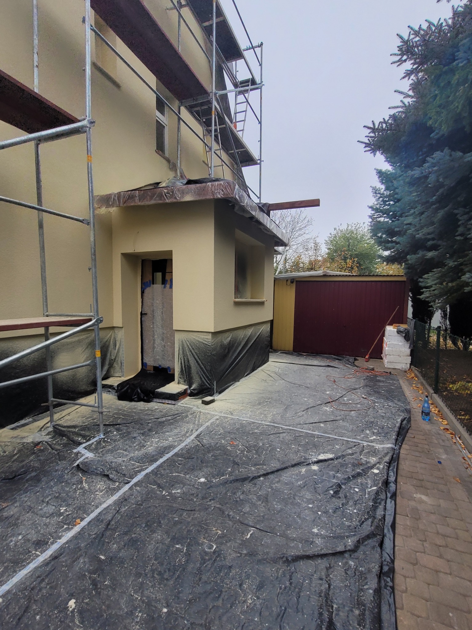 Dom jednorodzinny w trakcie prac elewacyjnych, widoczne rusztowania, zabezpieczona folią podstawa budynku i teren wokół, styropianowe płyty izolacyjne obok garażu.