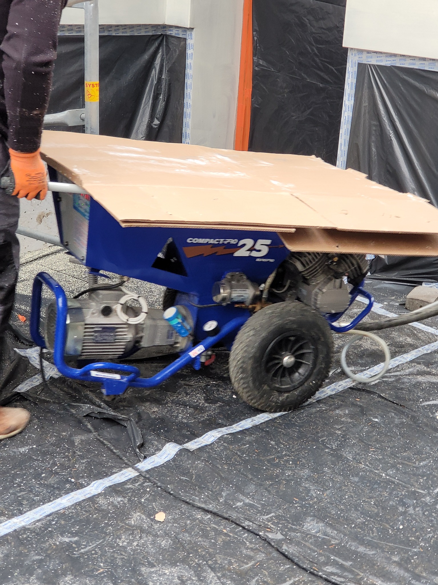 Niebieska maszyna do natrysku z silnikiem i kołami, częściowo zakryta kartonem, obsługiwana przez osobę w pomarańczowych rękawicach na folii ochronnej.