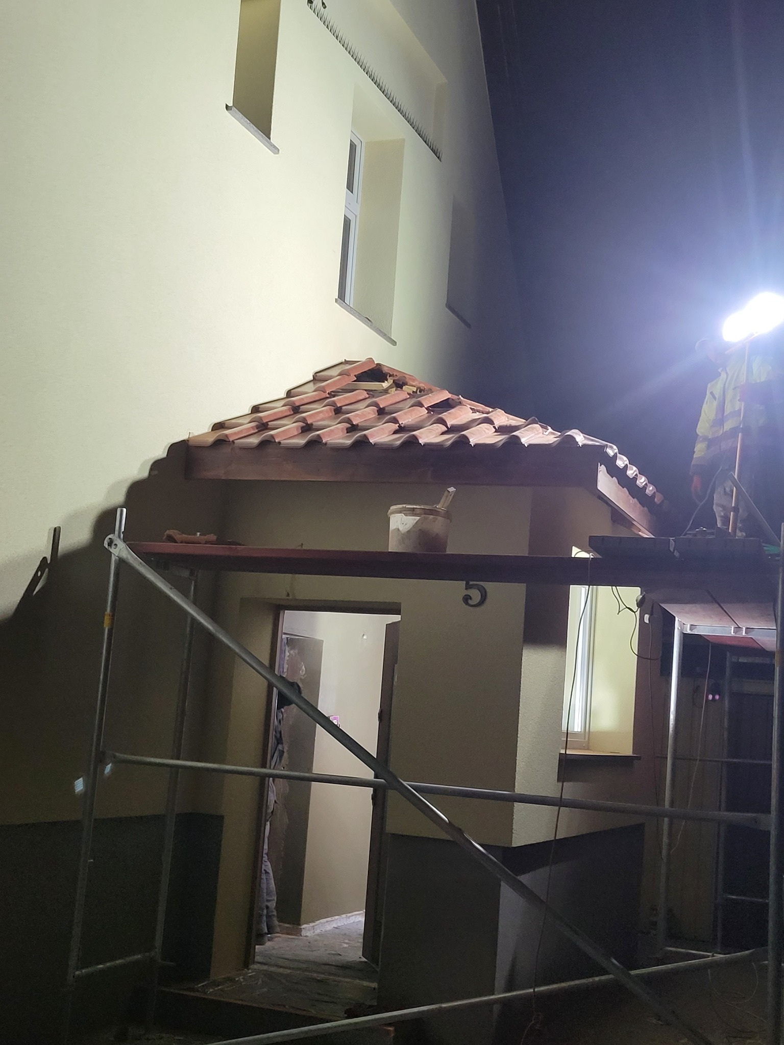 Nocna naprawa dachu nad wejściem do budynku z numerem 5, widoczne rusztowanie, wiadro z zaprawą i pracownik w odblaskowej kamizelce oświetlający miejsce pracy lampą.
